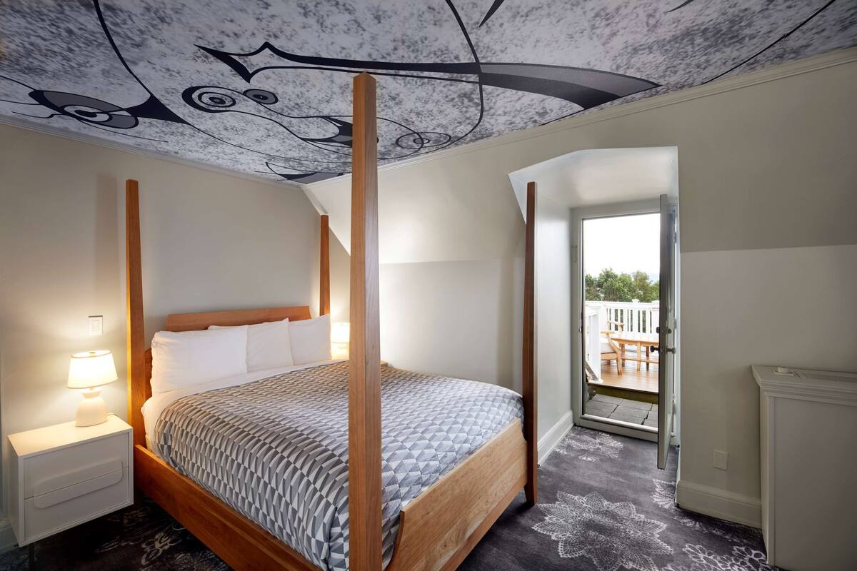 Een hemelbed met een deken met een geometrisch patroon, een nachtkastje met een lamp en een plafond met een uniek ontwerp.