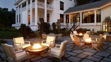 A large white building with columns and a balcony, outdoor seating with chairs and a fire pit, and a well-maintained garden.
