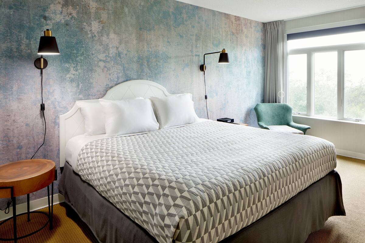 Een verzorgde hotelkamer met een groot bed, twee bedlampen, een houten nachtkastje en uitzicht op het omliggende landschap door een raam.