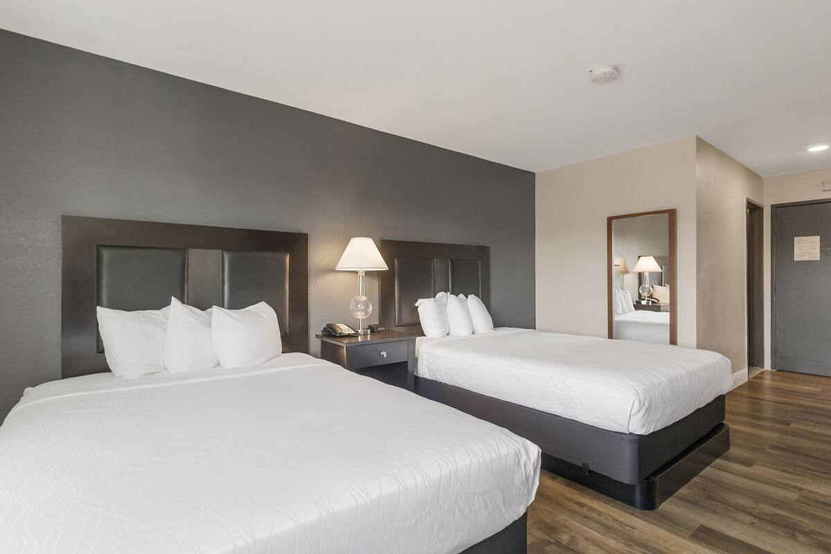 Quarto de hotel com duas camas, uma mesa de cabeceira com abajur, um espelho e uma porta.
