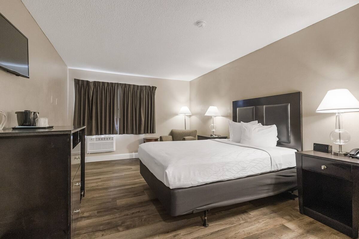 Quarto de hotel com cama, mesas de cabeceira, luminárias, TV de tela plana, escrivaninha com computador e janela com cortinas.