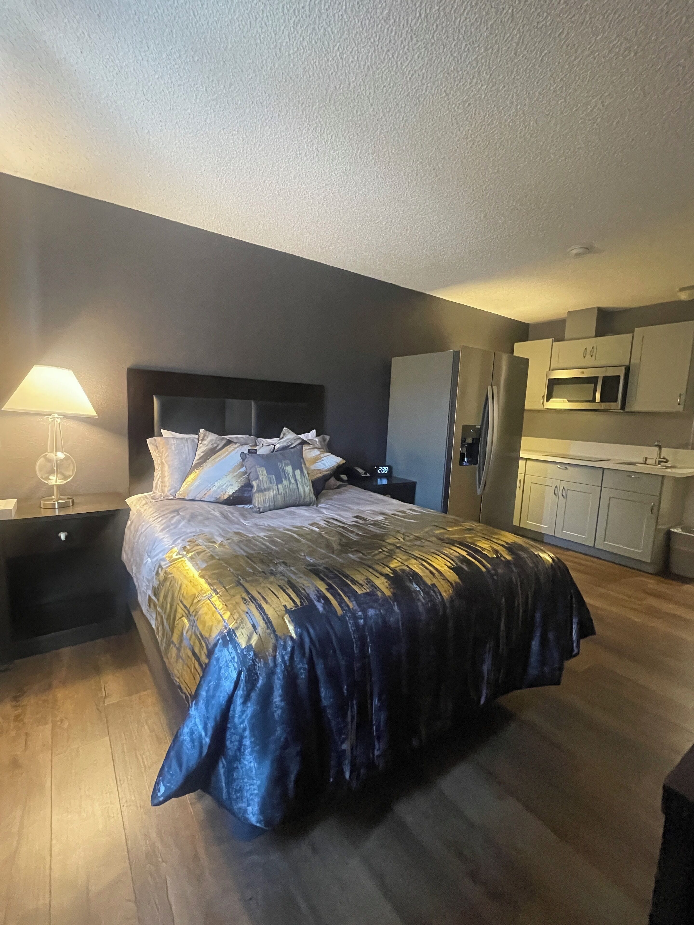 A bedroom with a bed, a nightstand, a lamp, a wall clock, and a kitchenette with cabinets and a microwave.