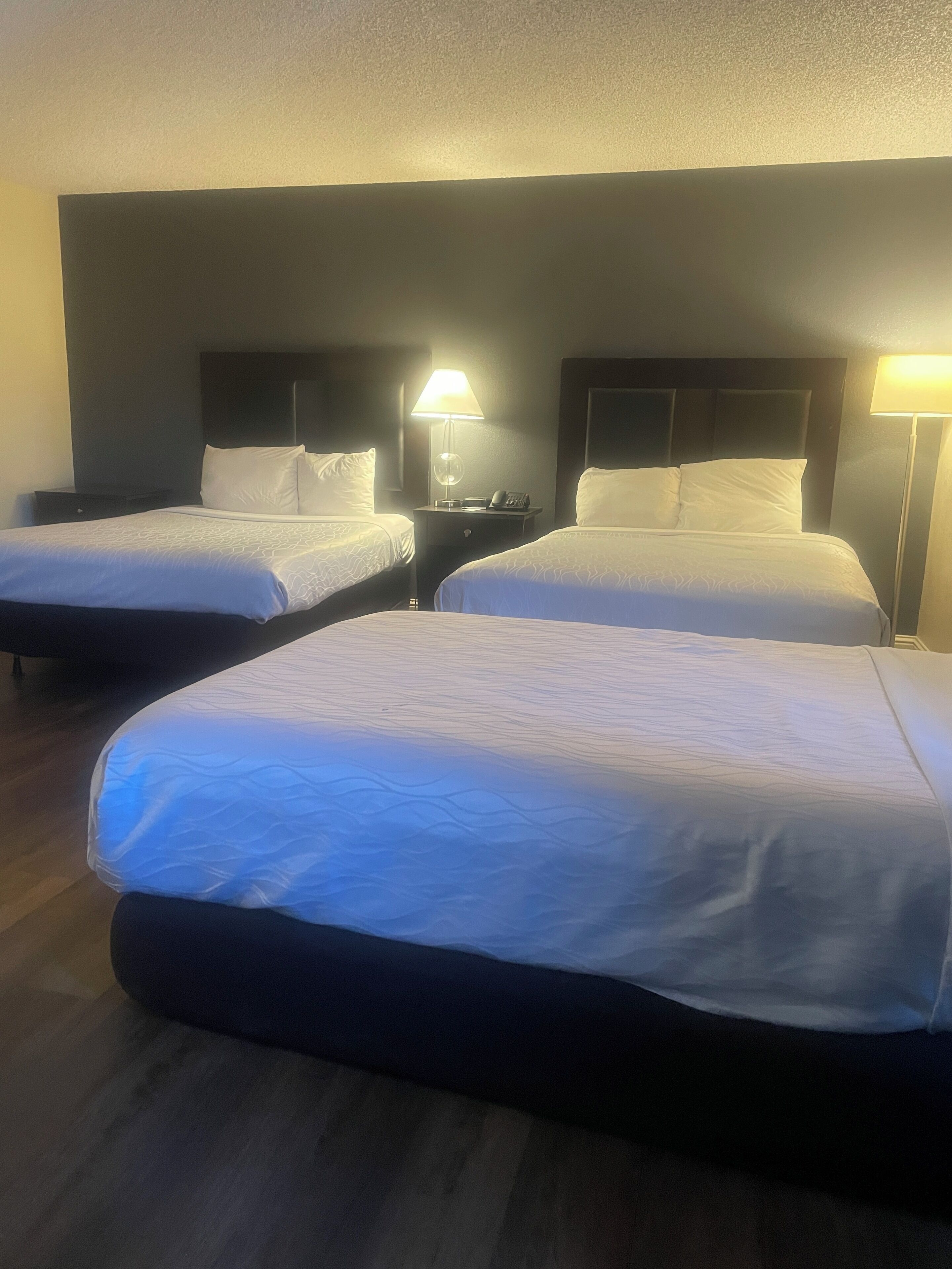 Camera d'albergo con due letti singoli e un letto più grande, tutti con lenzuola bianche. Sono presenti comodini con lampade e un tavolino tra i letti.