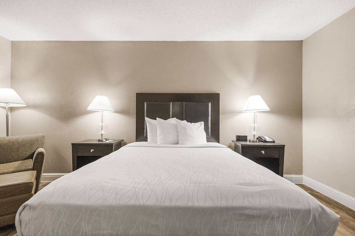 Quarto de hotel com uma cama grande, duas mesas de cabeceira com abajures, uma cadeira e uma cabeceira escura.