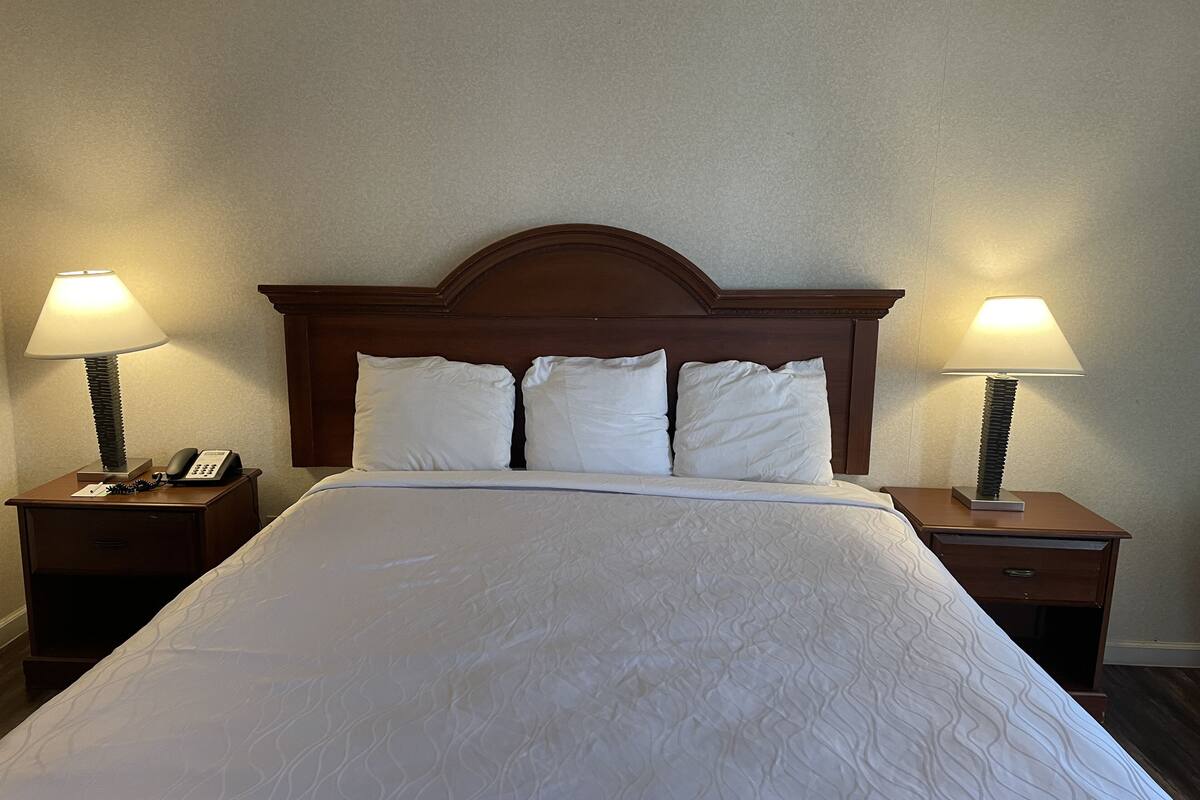 Cama bem arrumada com cabeceira de madeira, duas mesas de cabeceira com luminárias e um telefone na mesa da esquerda.