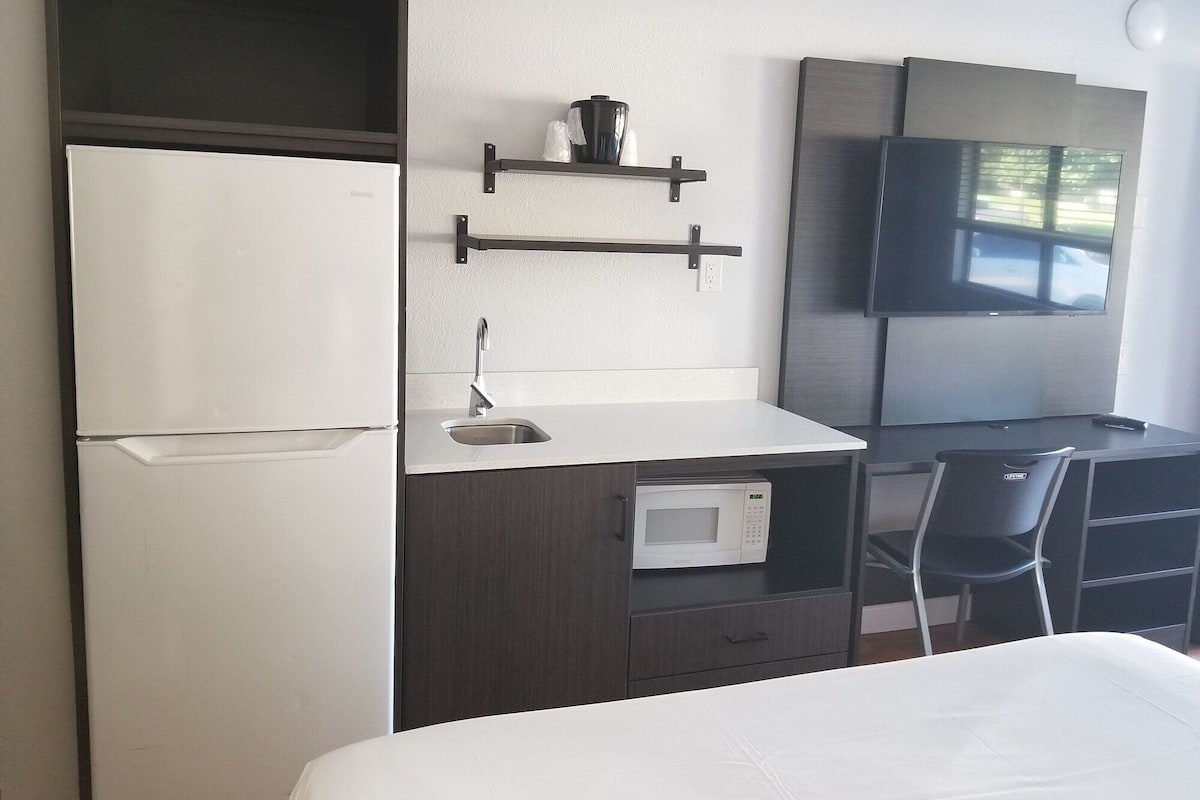 A hotel room with a bed, a desk with a microwave, a refrigerator, a TV, and a shelf with a coffee maker.