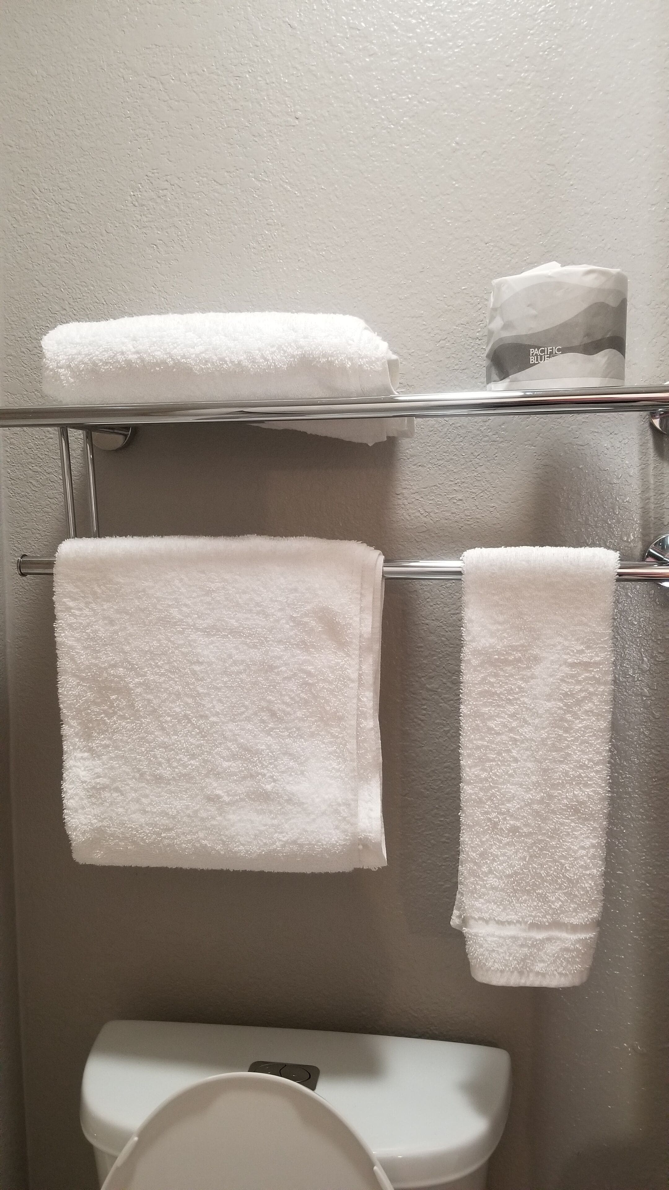 Un baño con dos toallas blancas colgadas en un perchero metálico, un inodoro y un dispensador de servilletas de papel con la etiqueta 'PACIFIC BLUE'.