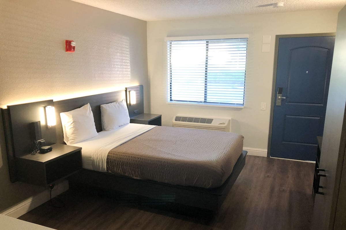 A hotel room with a bed, bedside tables, a window with blinds, an air conditioning unit, and a door.
