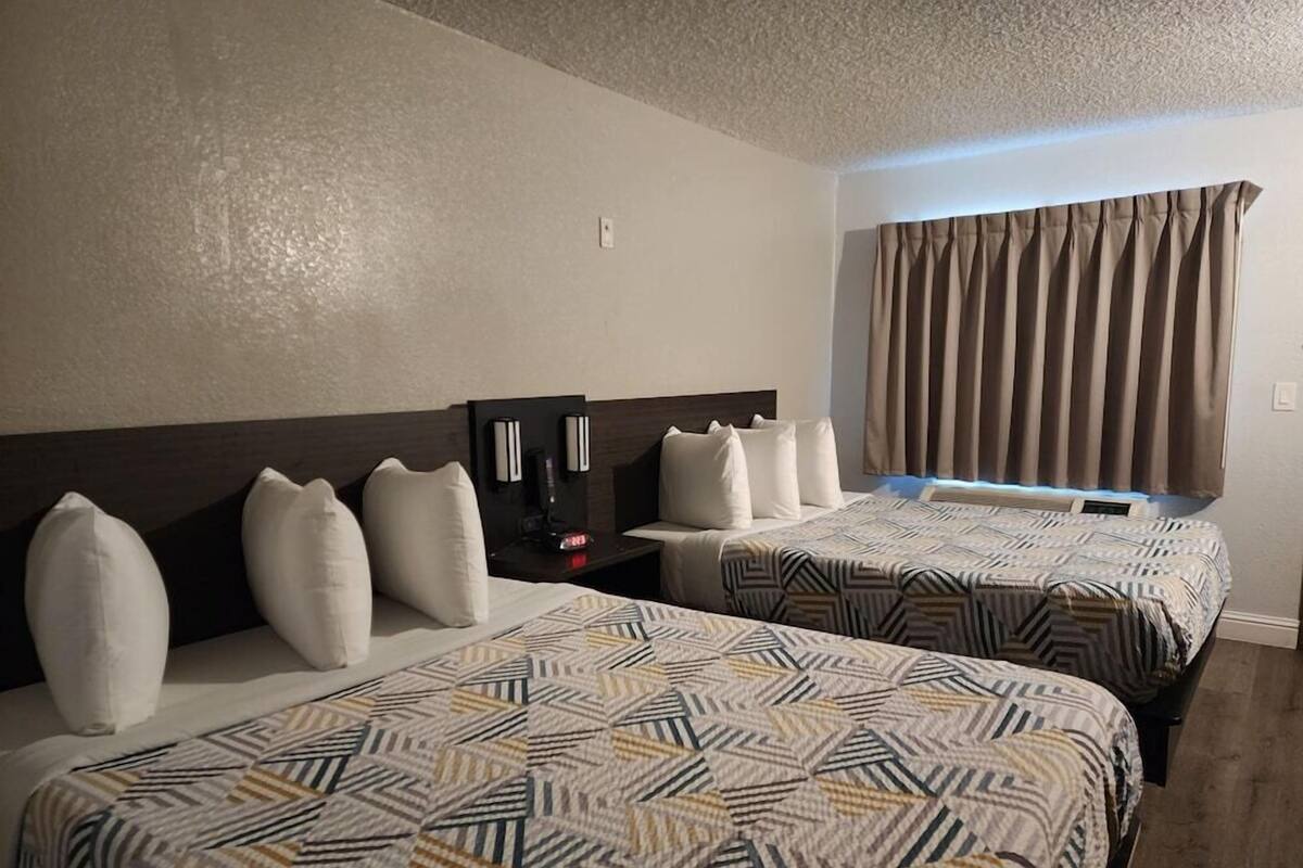 A hotel room with two beds, a headboard, a nightstand, a clock, and a window with curtains.