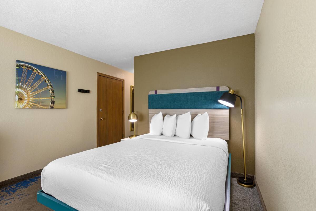 A hotel room with a bed, a headboard, a wall-mounted lamp, a door, and a wall painting.