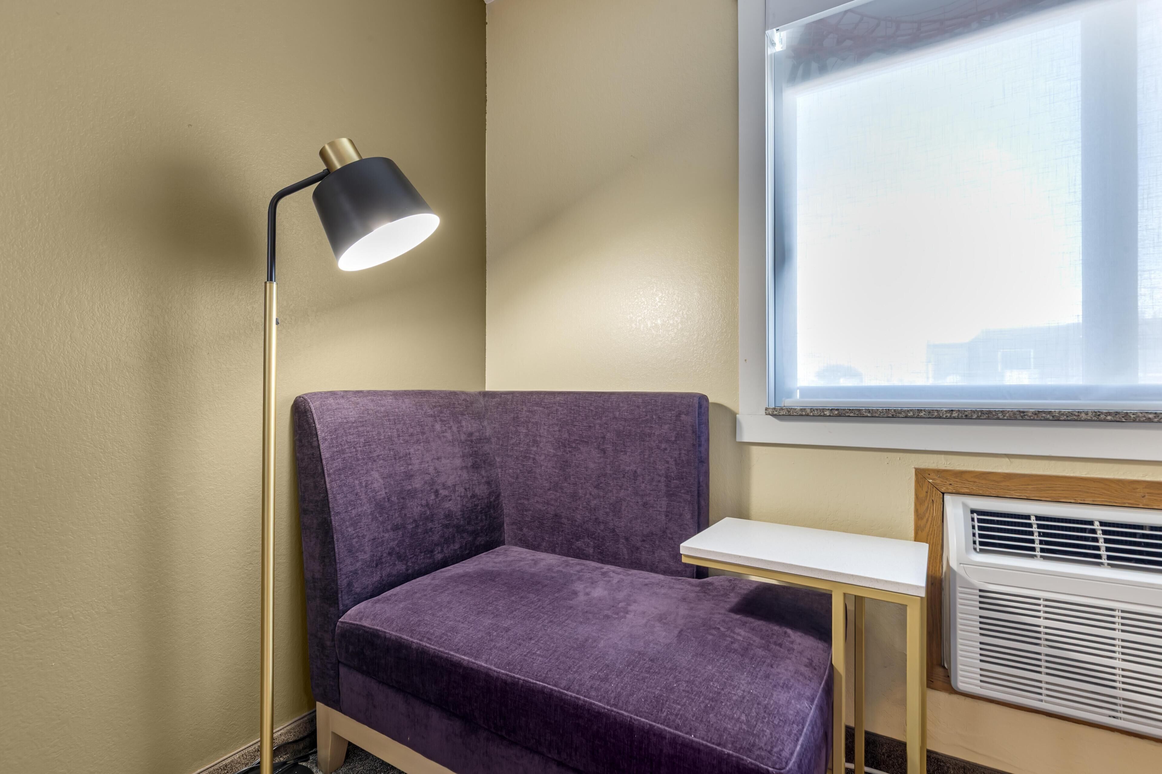 Une pièce avec un canapé violet, un lampadaire, une petite table et une fenêtre avec des stores.