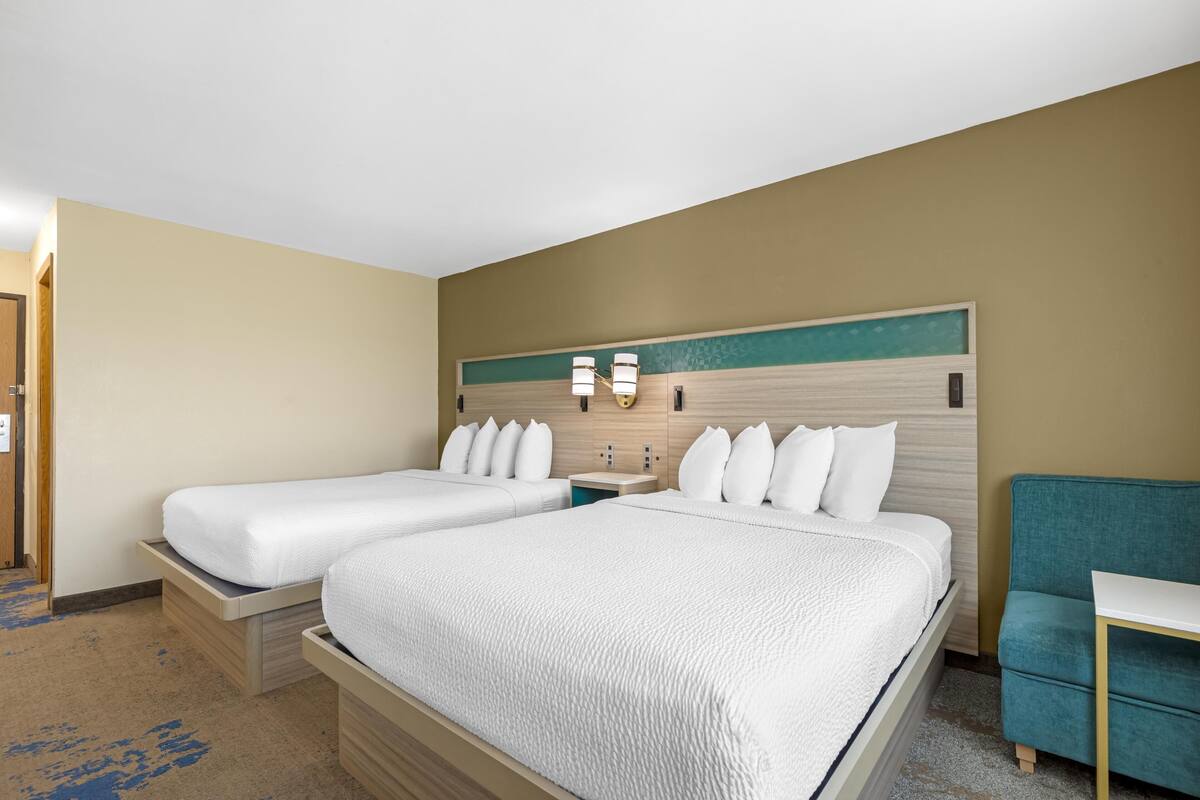 A hotel room with two beds, a bedside table, a nightstand, and a teal armchair.