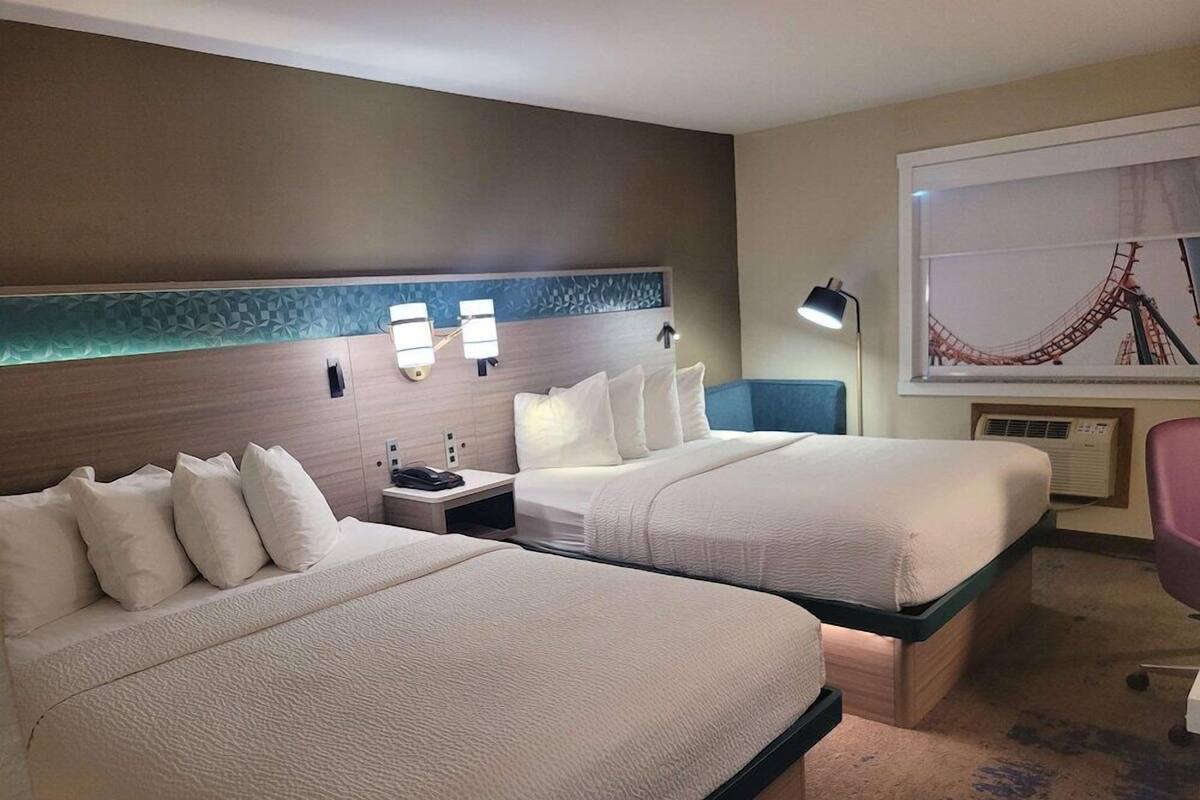 A hotel room with two beds, a nightstand, a desk, a chair, and a large window with a view of a roller coaster.