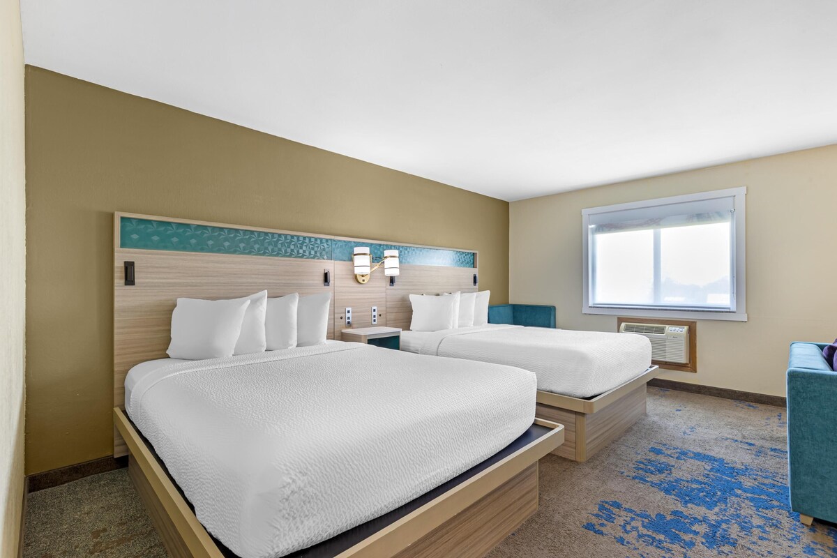 Een hotelkamer met twee bedden, een groot raam, een blauw tapijt en een blauwgroene stoel.