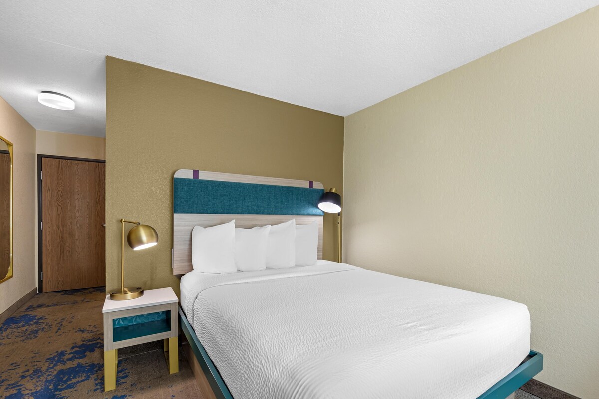 Een hotelkamer met een bed, nachtkastje, lamp en een deur.