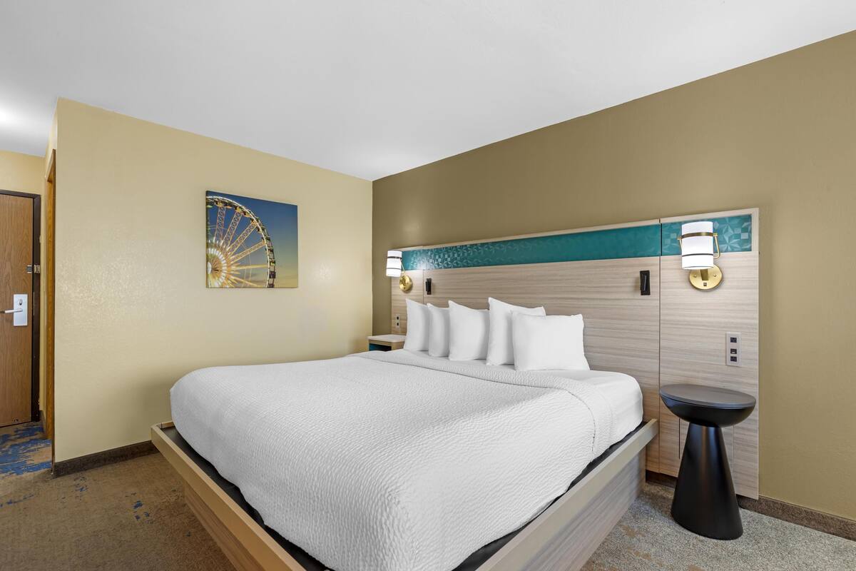 A hotel room with a bed, a headboard with wall-mounted lamps, a small side table, and a wall-mounted artwork of a Ferris wheel.