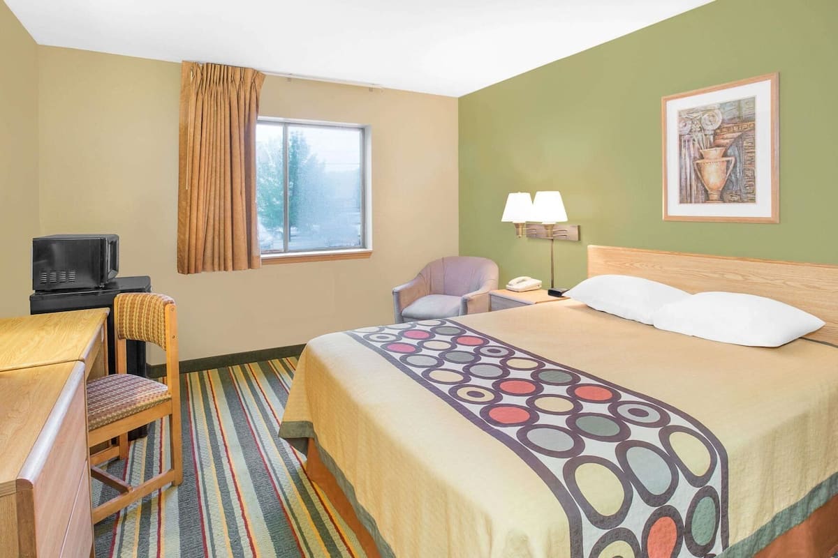 Une chambre d’hôtel comprenant un lit, un bureau, une chaise, une télévision, une fenêtre avec des rideaux, une lampe et un tableau au mur.