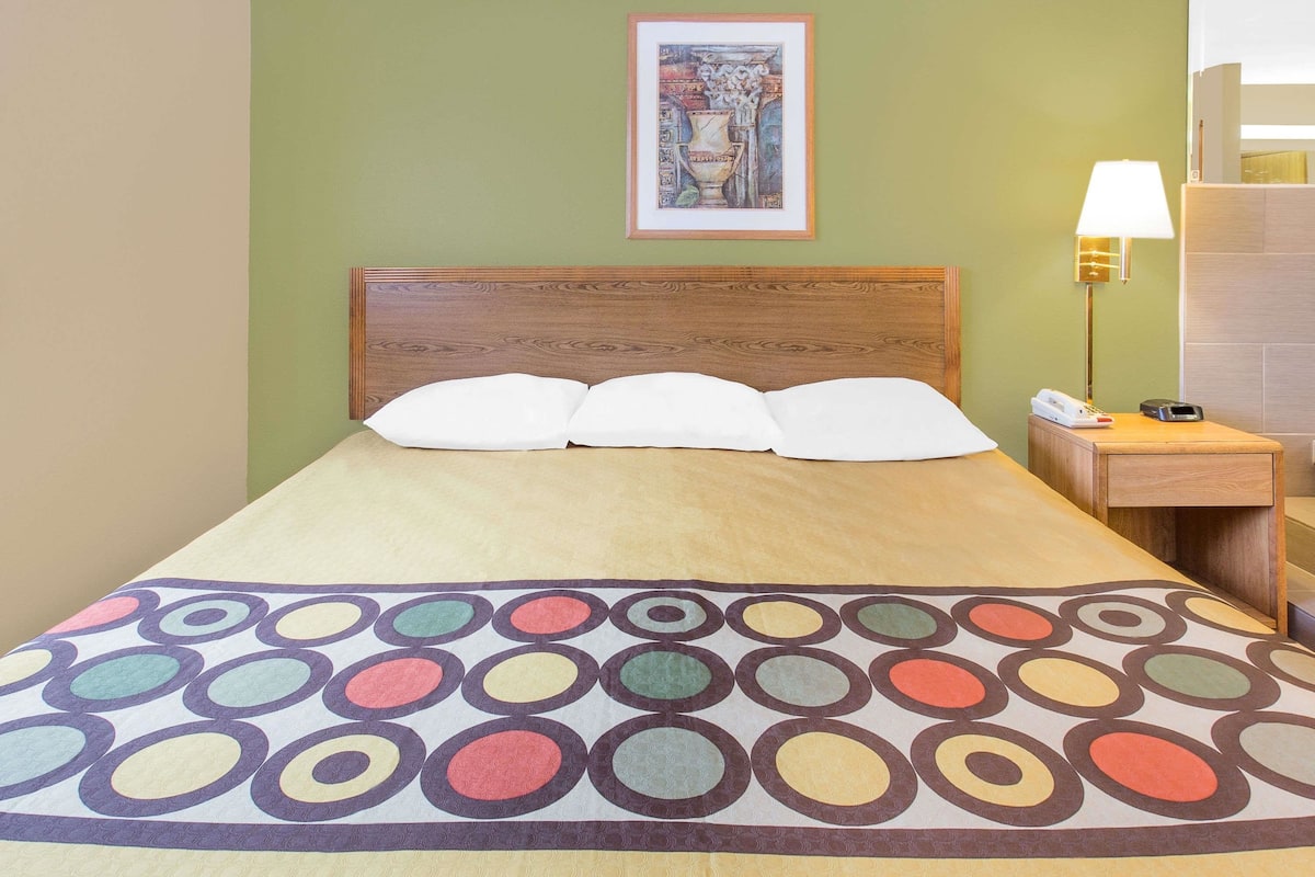 Un lit bien fait, recouvert d’une couvre-lit aux motifs circulaires et colorés, avec une tête de lit en bois, une lampe de chevet et un tableau encadré accroché au mur.