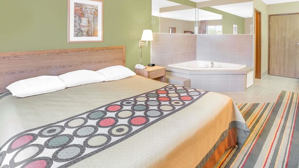 A hotel room with a bed, a bedside table with a lamp, a large mirror, a bathroom with a bathtub, and a door. - Super 8 by Wyndham East Moline (East Moline)