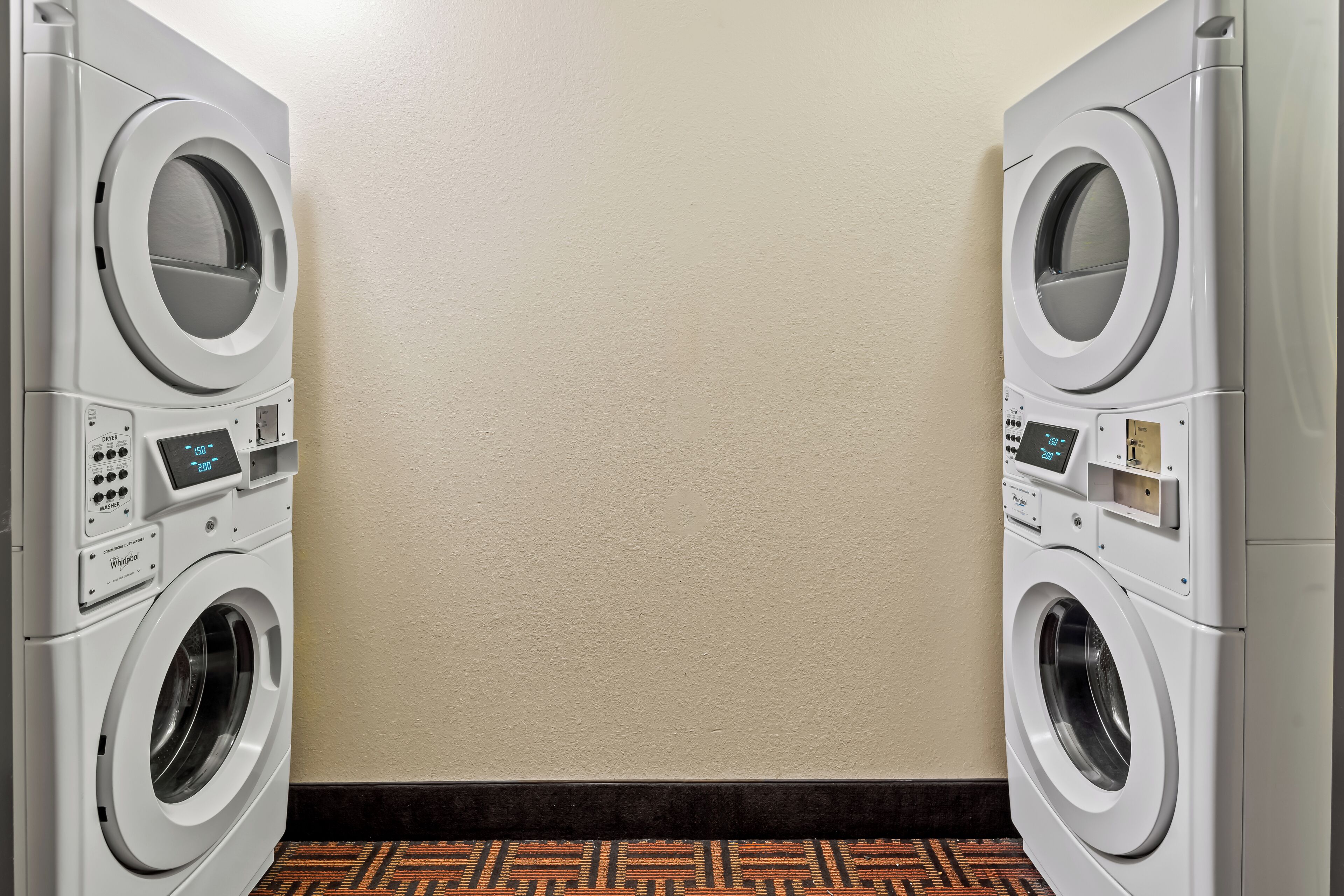Deux lave-linge Whirlpool à chargement frontal sont installés dans une buanderie, avec un mur uni et un sol à motifs.