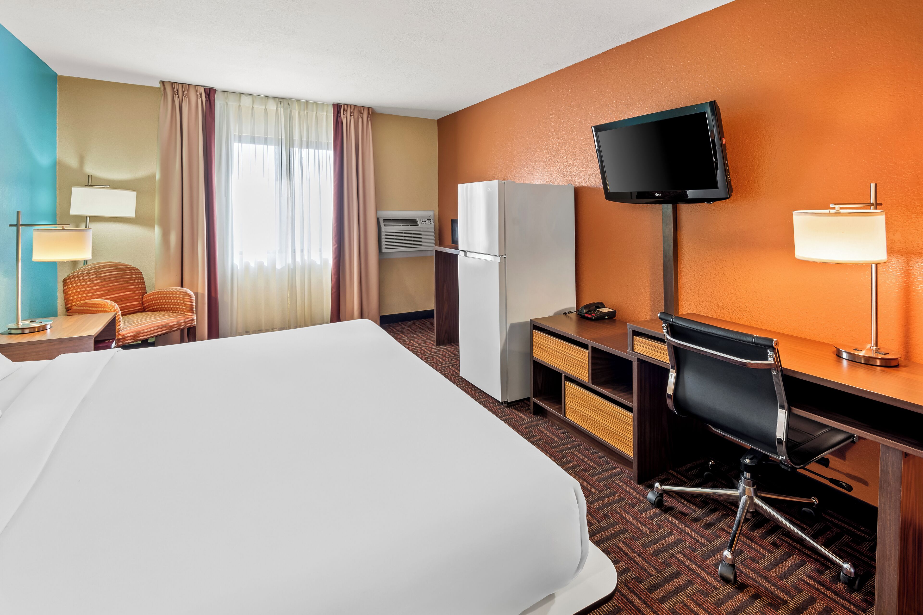 Habitación de hotel con una cama grande, un escritorio con computadora, una silla, televisor montado en la pared y un refrigerador.