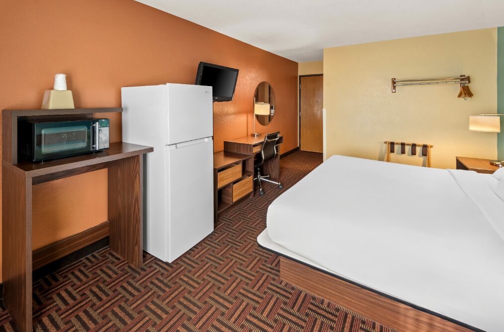 A hotel room with a bed, a desk with a computer, a microwave, a refrigerator, a TV, and a lamp.