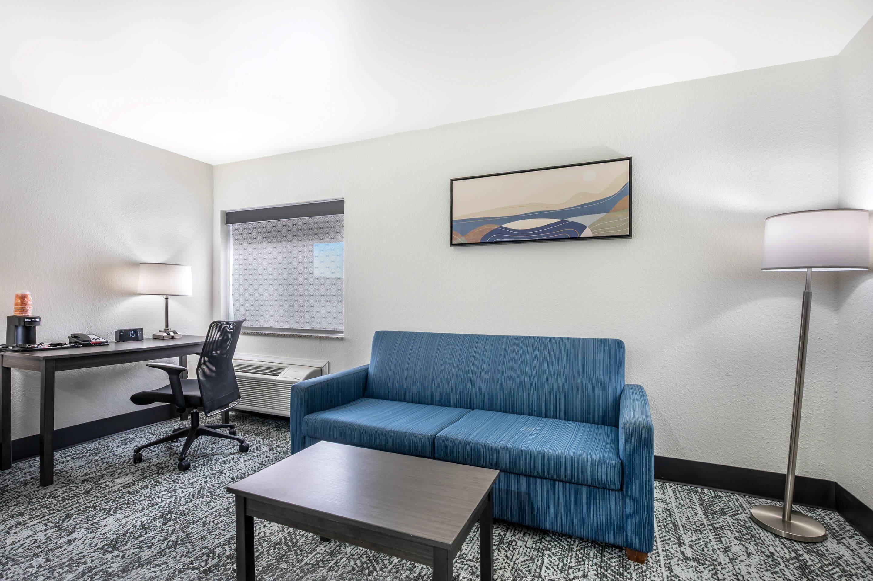 A hotel room with a blue sofa, a desk with a chair, a coffee maker, a lamp, and a painting on the wall.