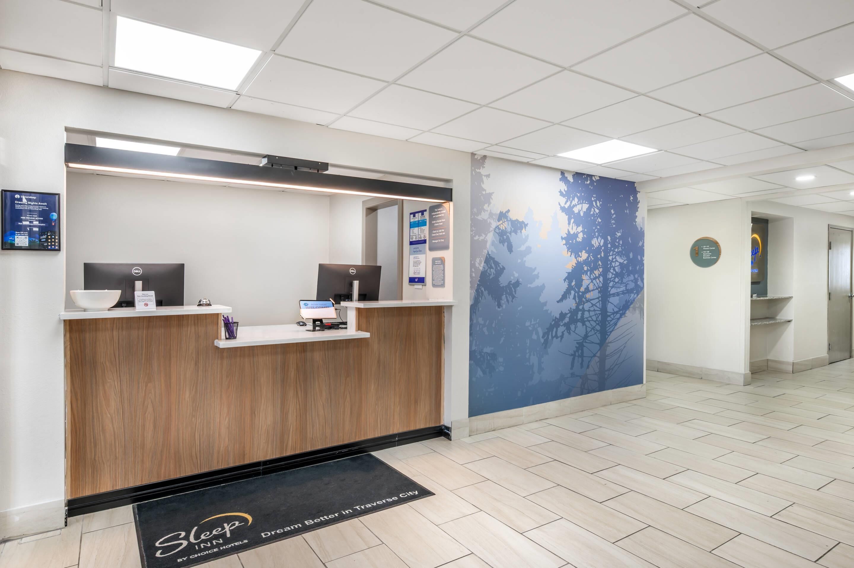 A reception area with a wooden reception desk, two computer monitors, a digital display, and a wall with a tree design.
