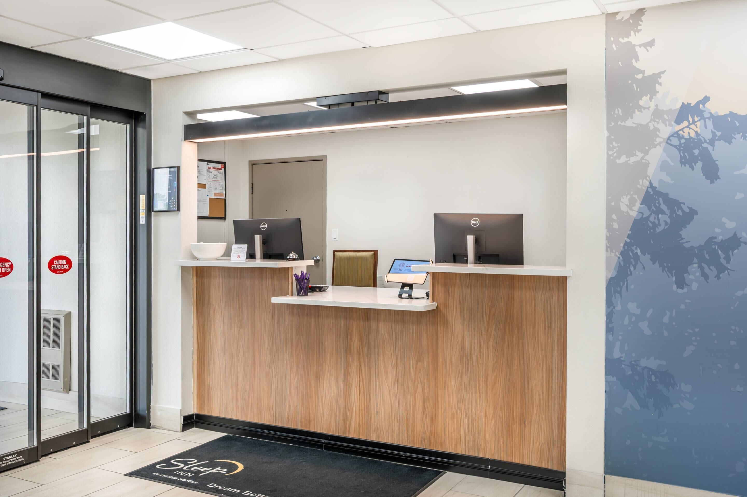A hotel reception desk with two computer monitors, a small shelf with pens, and a digital screen.