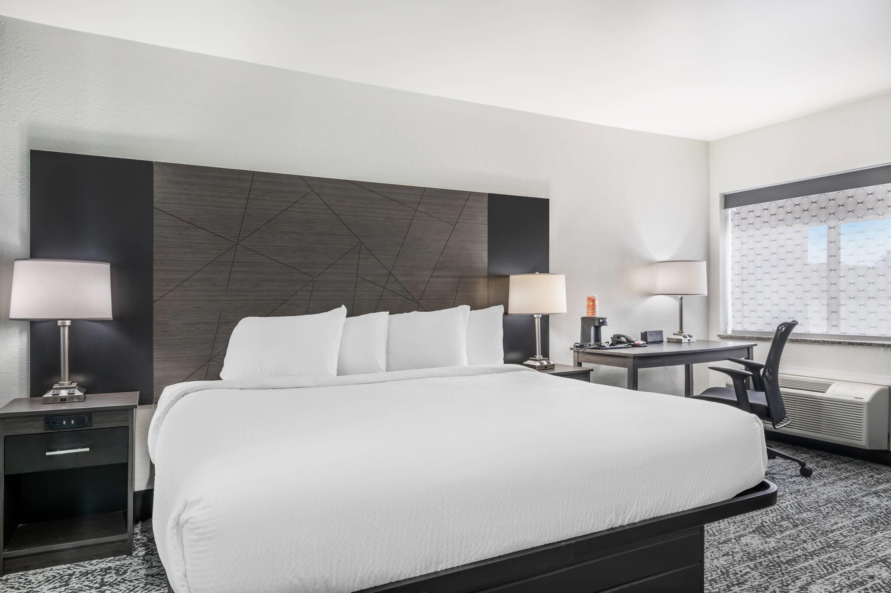 A hotel room with a large bed, two bedside tables with lamps, a desk with a chair, and a window with a view.