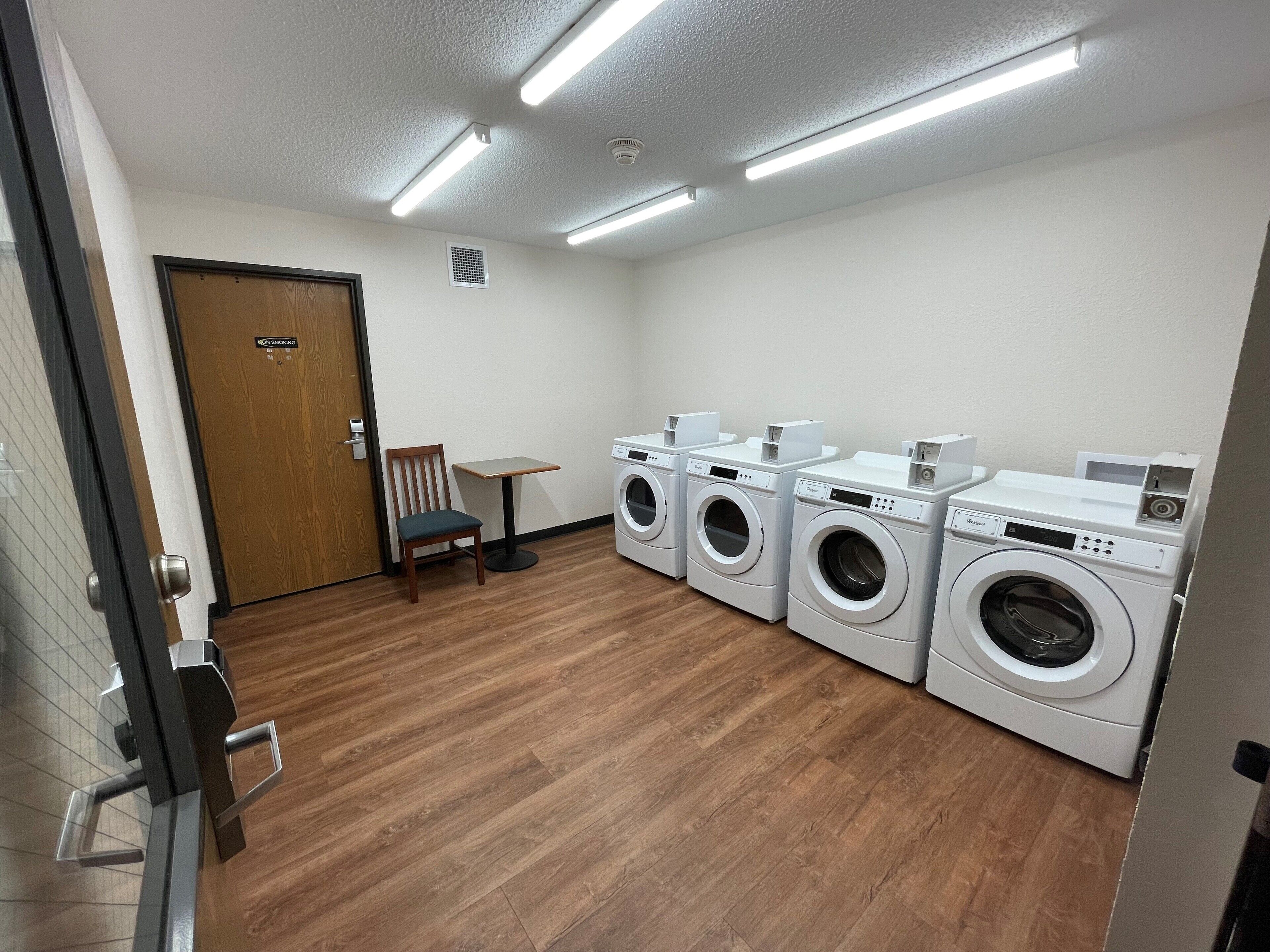 A laundry room with four washing machines, a wooden chair, a small table, and a door with a sign.