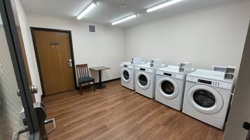 A laundry room with four washing machines, a wooden chair, a small table, and a door with a sign.
