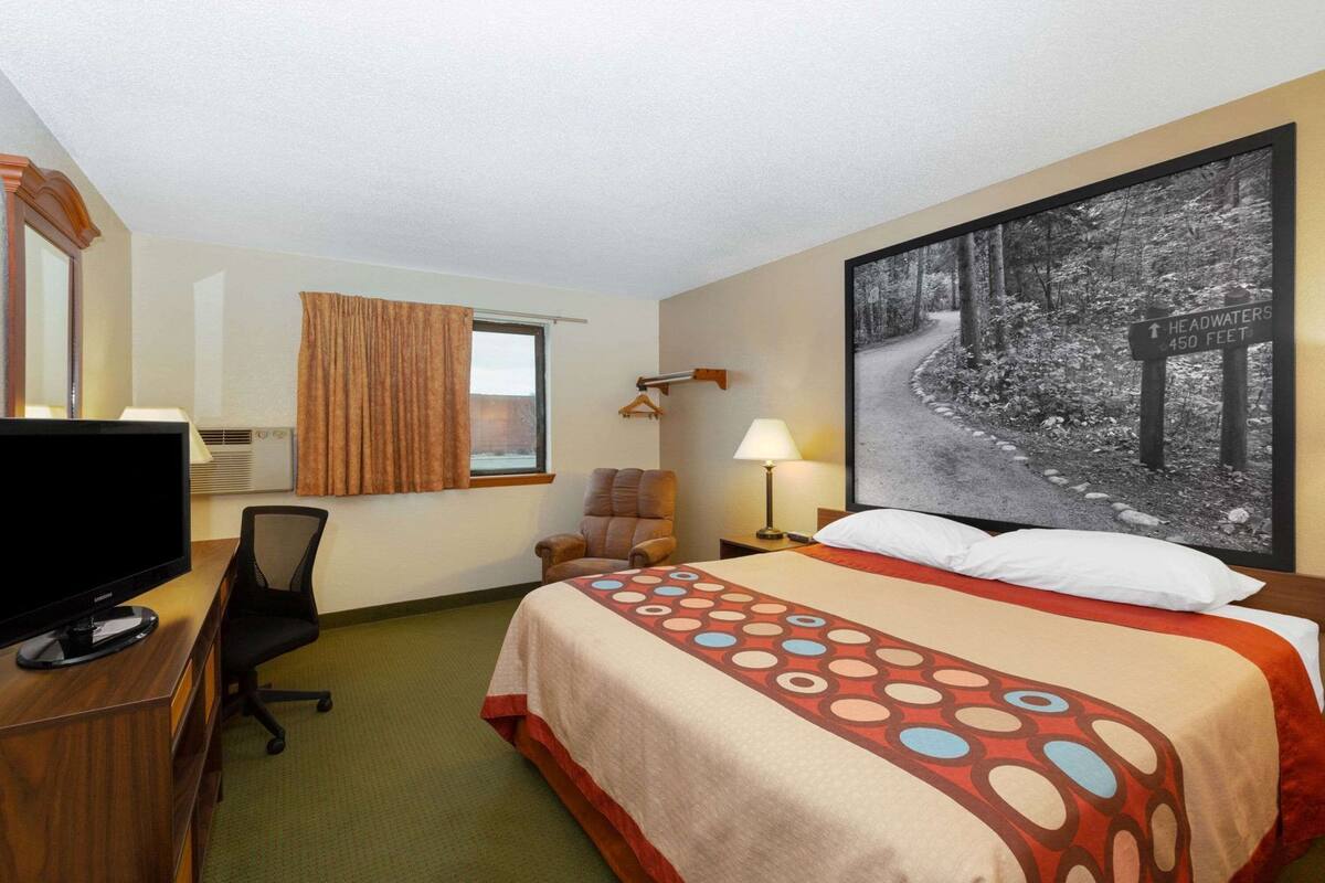 Une chambre d’hôtel avec un lit, un bureau avec un ordinateur, une chaise, une lampe et un grand tableau encadré au mur.