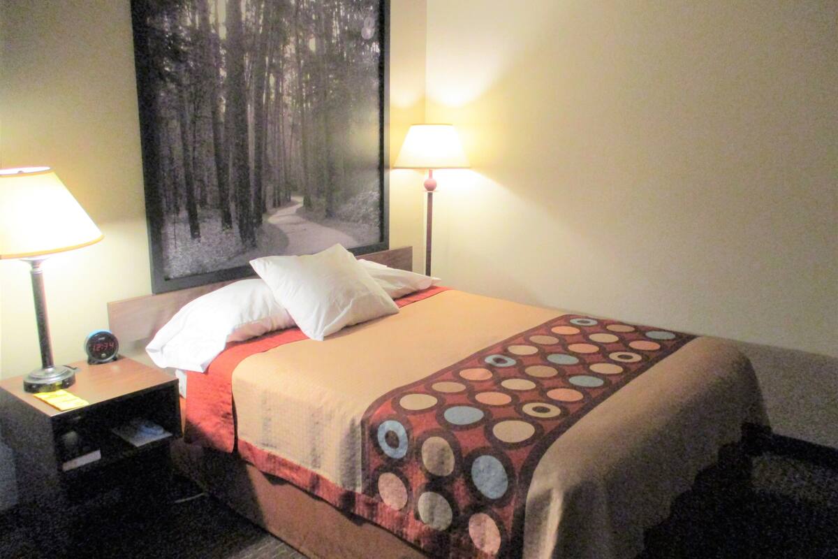 Une chambre d’hôtel avec un lit, deux lampes de chevet, une table de nuit avec une horloge et un grand tableau au mur.