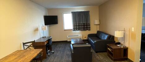 A hotel room with a wooden desk, a black leather sofa, a small table, and a TV mounted on the wall.