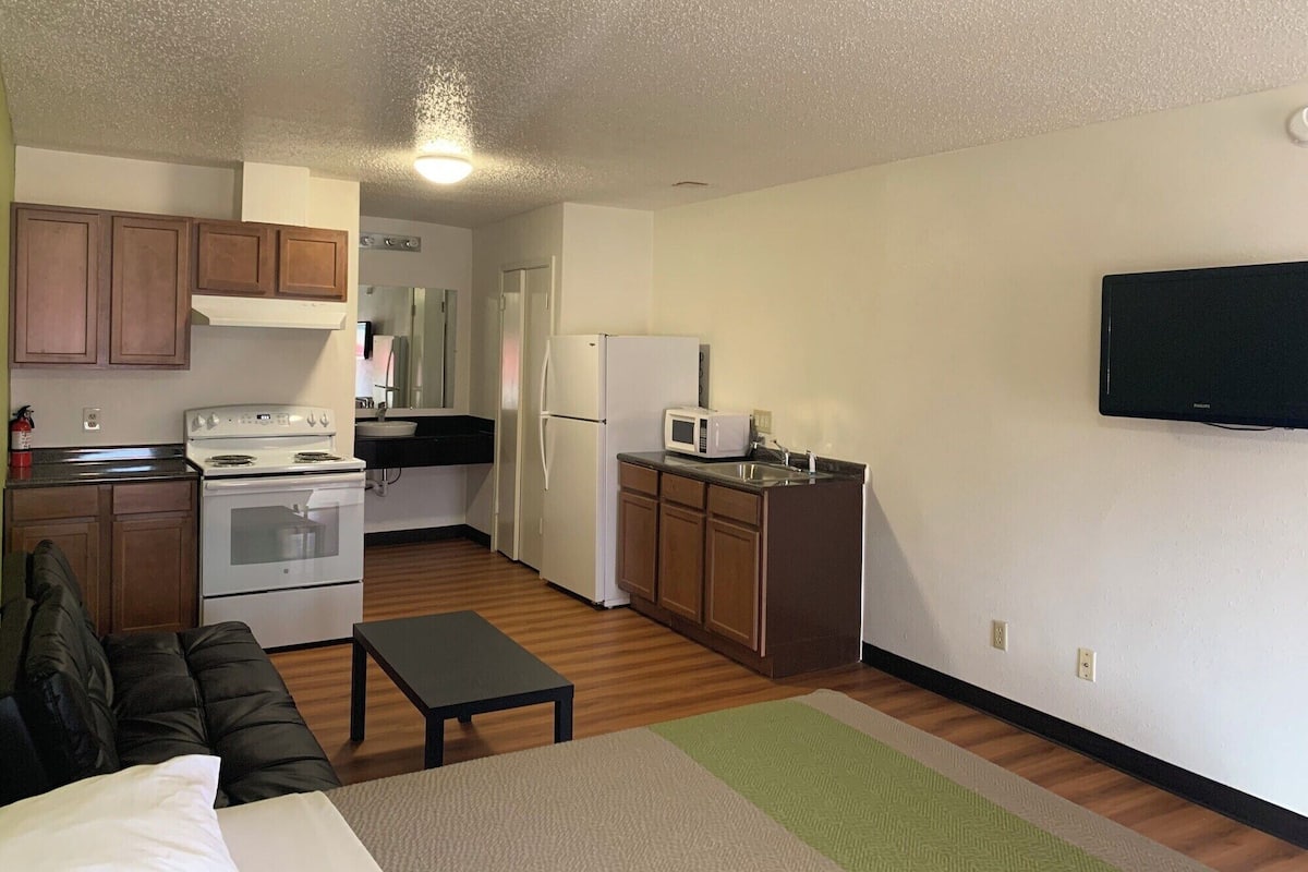 A compact kitchen with a stove, microwave, and refrigerator. There's a small dining area with a table and a couch. A flat-screen TV is mounted on the wall.