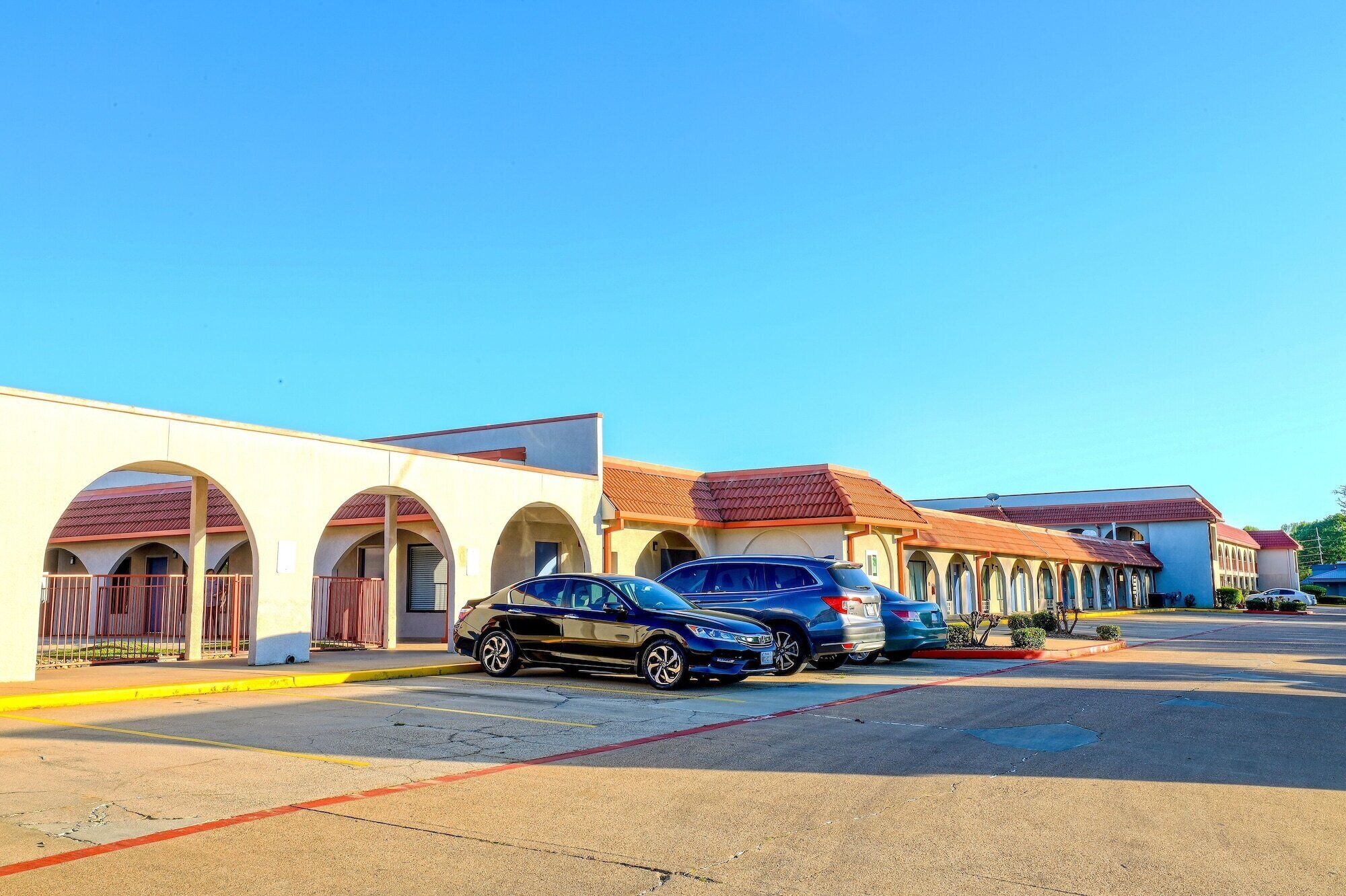 A parking lot with several cars parked in front of a building with arched windows and a red roof.