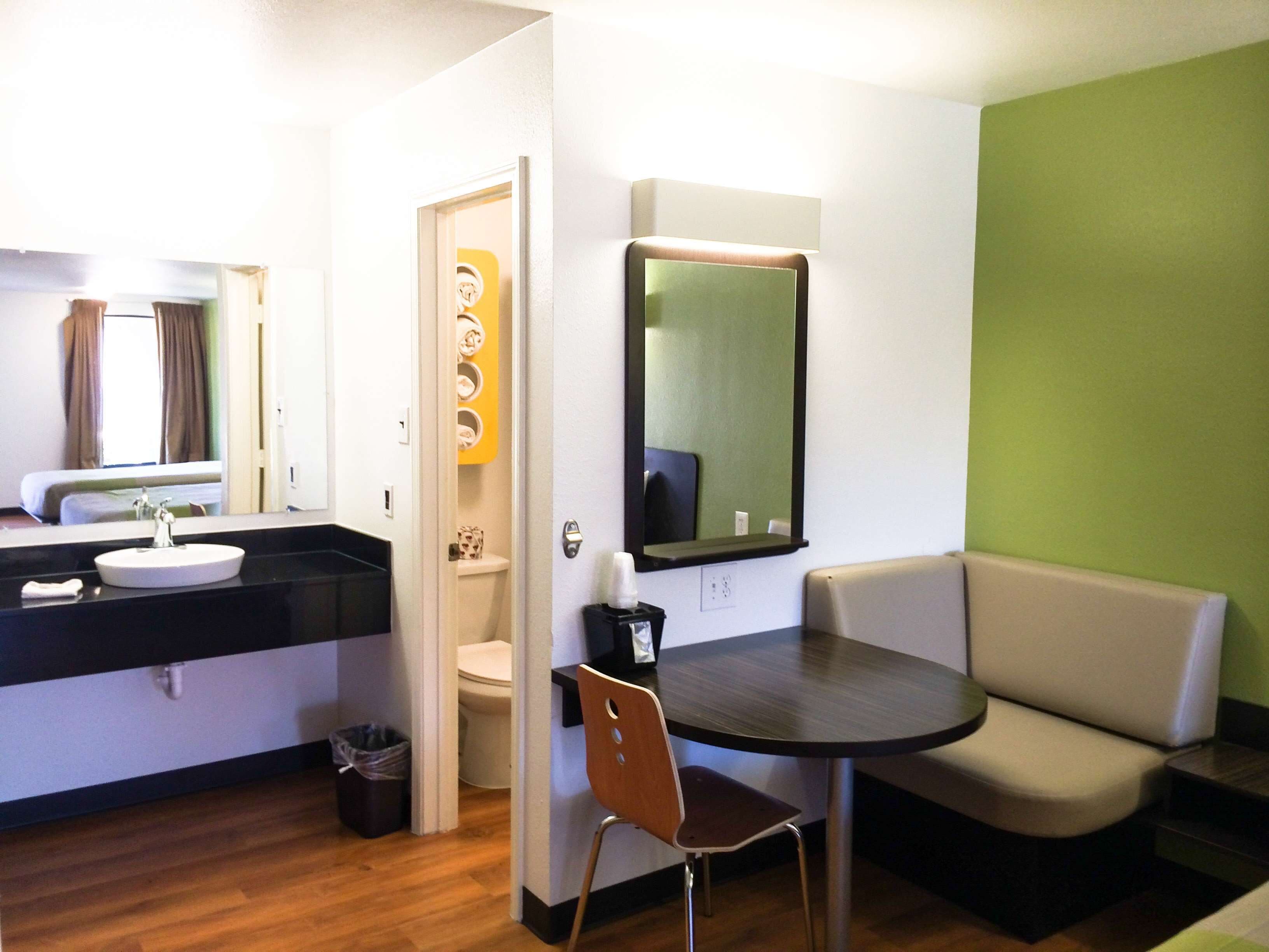 A hotel room with a round dining table, a sofa, a small chair, a bathroom with a toilet and sink, and a mirror on the wall.