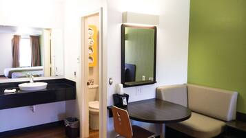 A hotel room with a round dining table, a sofa, a small chair, a bathroom with a toilet and sink, and a mirror on the wall.