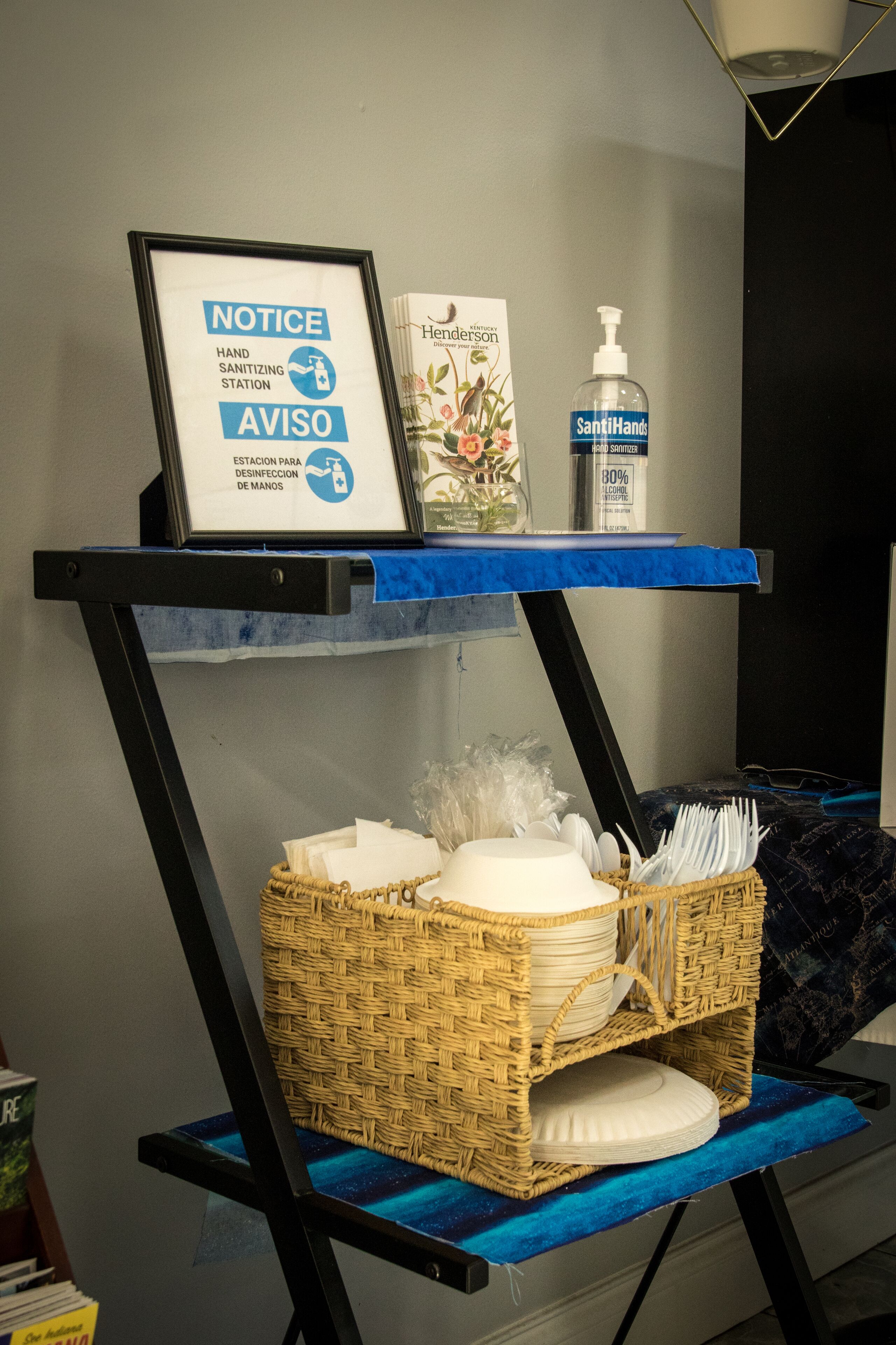 A hand sanitizing station with a notice, a SantiHands bottle, and a basket with plates and napkins on a blue shelf.