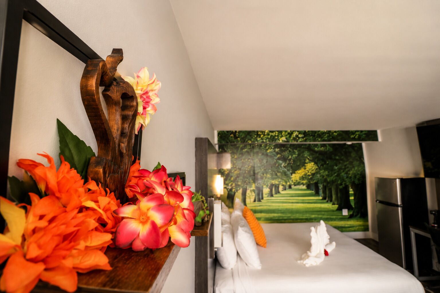 A hotel room with a bed, a wooden sculpture, and a painting of a tree-lined path.