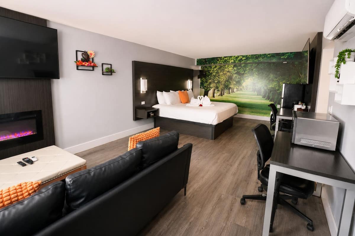 A modern hotel room with a bed, a desk, a sofa, a fireplace, and a large window with a forest view.