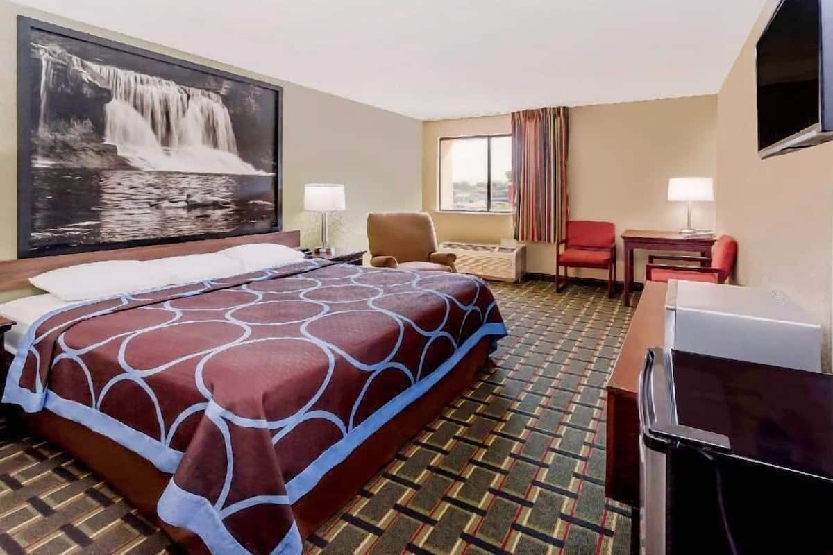 A hotel room with a large bed, a desk, a chair, a TV, and a painting of a waterfall.