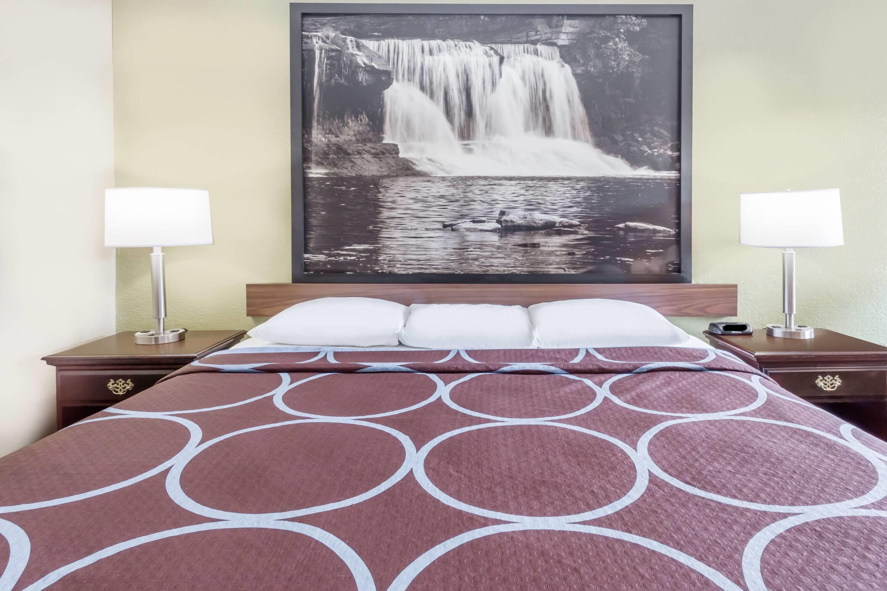 Quarto de hotel com cama, duas mesas de cabeceira, um criado-mudo, fotografia grande de uma cascata emoldurada na parede e duas luminárias.