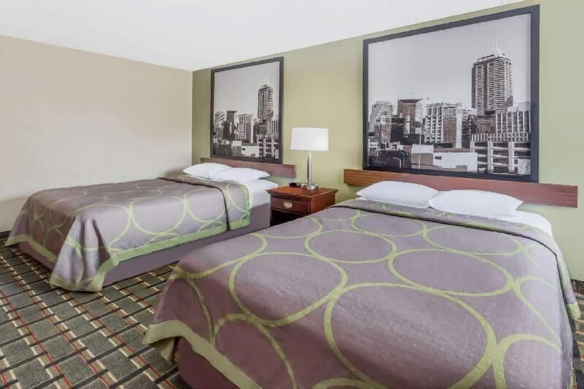 A hotel room with two beds, a nightstand, a lamp, and two cityscape wall art pieces.