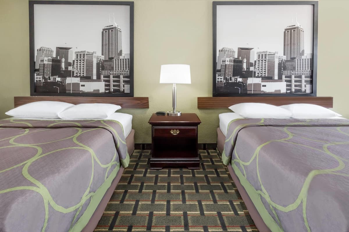 A hotel room with two beds, a nightstand with a lamp, and two cityscape posters on the wall.