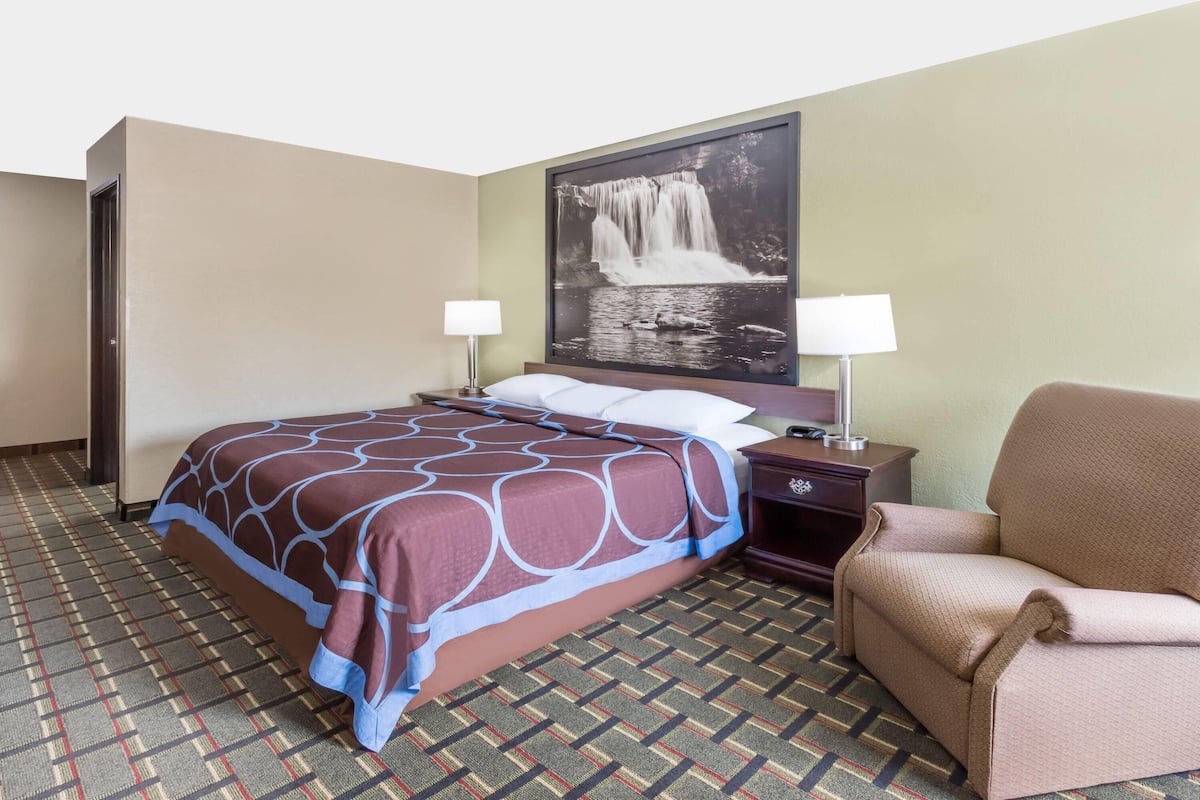 A hotel room with a large bed, a chair, a nightstand, a lamp, and a wall-mounted picture of a waterfall.