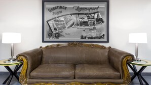 A brown leather sofa with ornate gold detailing, flanked by two small tables with lamps, against a wall with a framed black and white cityscape poster. - Super 8 by Wyndham Logansport (Logansport)