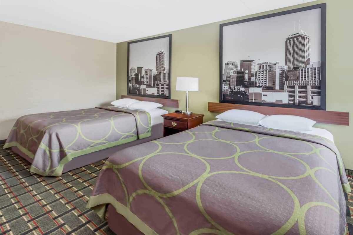 A hotel room with two beds, a nightstand, a lamp, and two framed cityscape pictures on the wall.