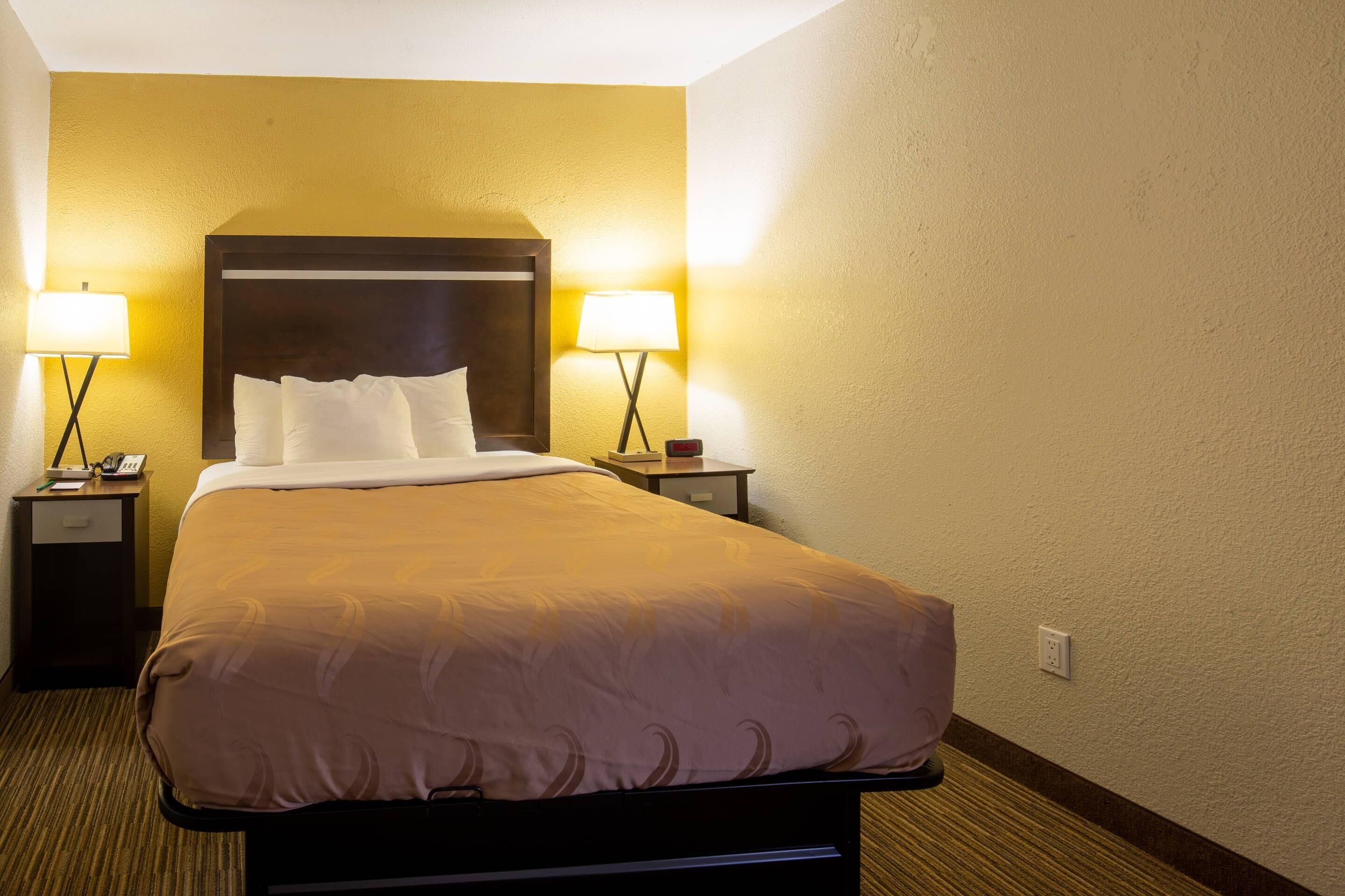 A hotel room with a bed, two bedside lamps, a nightstand with a phone, and a wall socket.